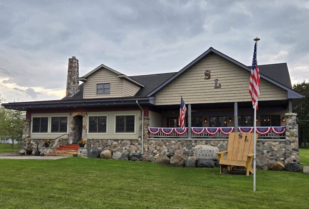Stone building with a chimney, decorated with American flags and red, white, and blue bunting. A large wooden chair and a sign reading "The Stone Lodge" are in front.