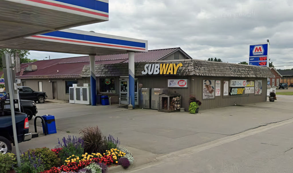 Gas station with a Subway restaurant, flower bed in the foreground, and gas prices displayed on a sign.