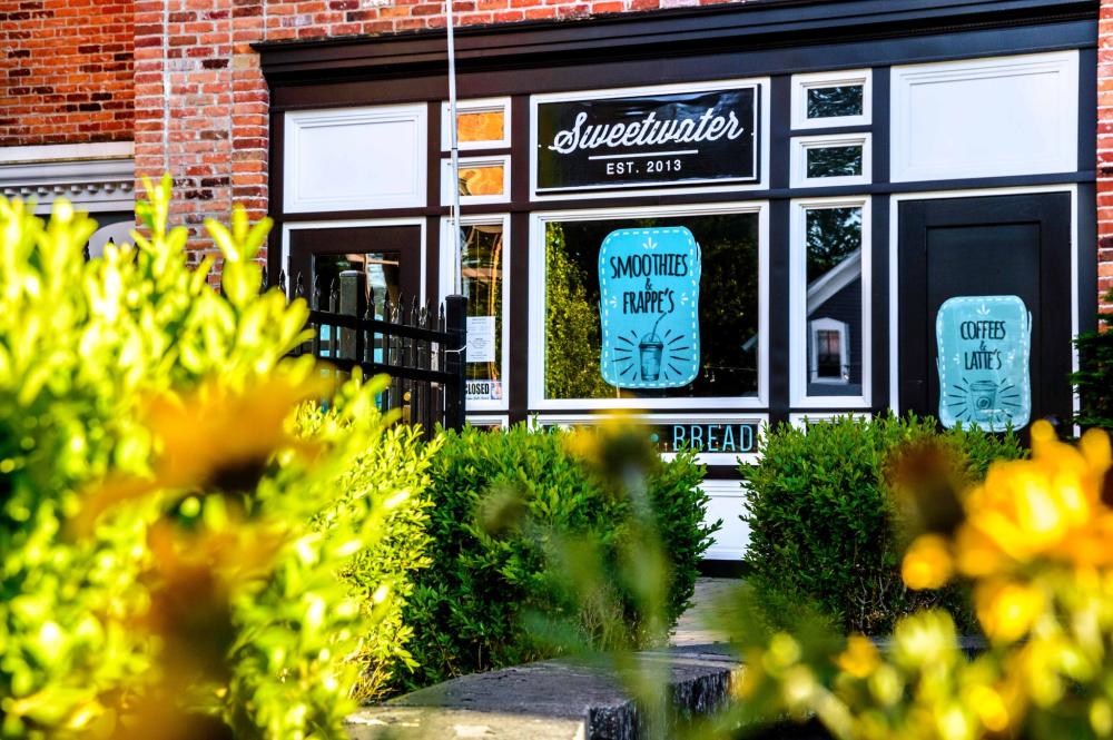 Front of a brick cafe named "Sweetwater" with signs for smoothies, frappés, coffees, and lattes, surrounded by green shrubs and yellow flowers in the foreground.