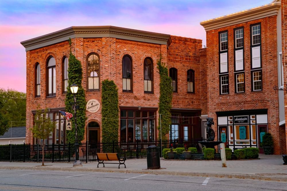 Historic brick building with large arched windows, ivy climbing the walls, and a sign reading "Ambassador." An American flag and a lamppost are in front, with a bench and a small statue nearby. The sky is pink and blue at sunset.