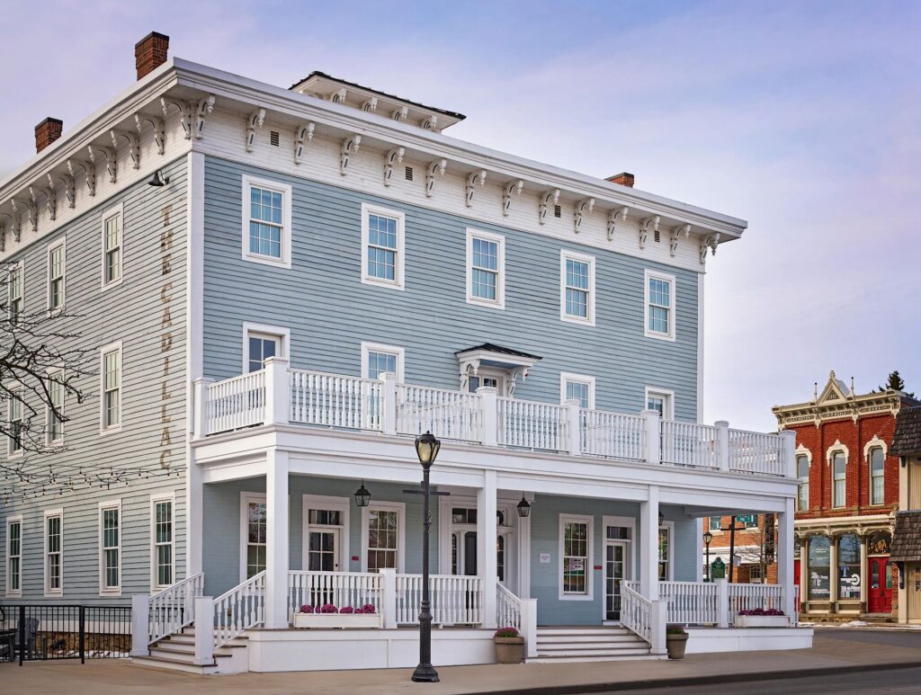 Historic three-story building with light blue siding, white trim, and a wraparound porch.