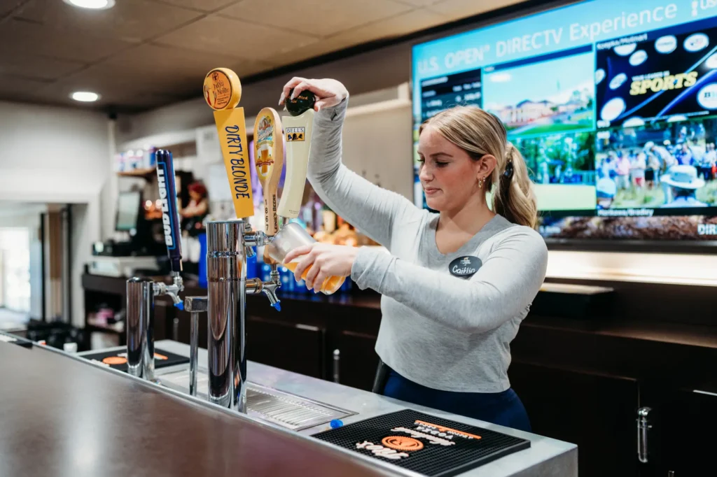 Bartender pouring a draft beer from a tap labeled "Dirty Blonde" at a bar with multiple screens in the background.