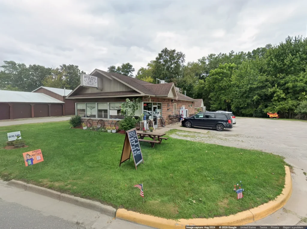 A small, rustic store with a brown roof and large windows, surrounded by greenery. A black vehicle is parked in the gravel lot. A picnic table and a chalkboard sign are on the grass in front. Trees are in the background.