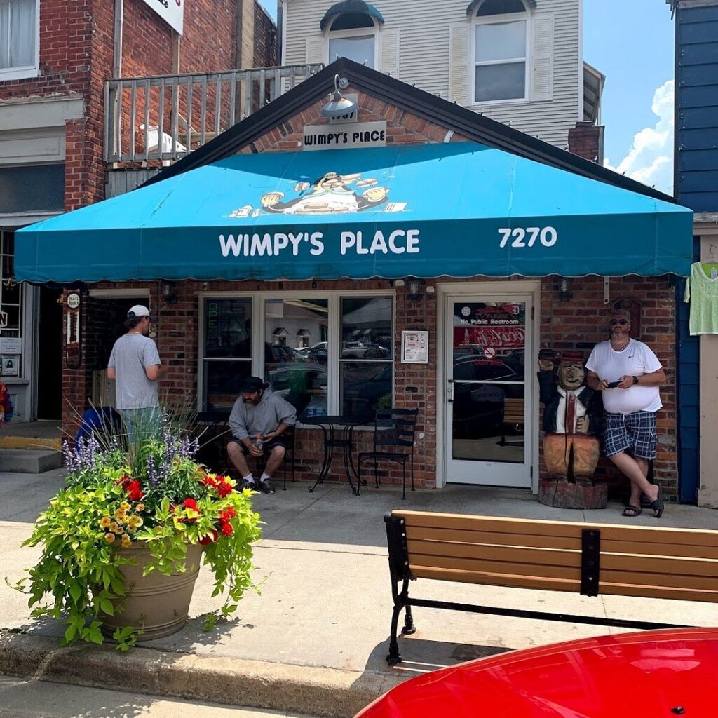 Front view of Wimpy's Place with a blue awning, a bench, and a flower planter outside. Three people are standing or sitting near the entrance.